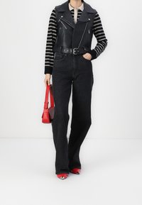 Person, der ein schwarz gestreiftes Langarmshirt, eine schwarze Lederweste, dunkle Jeans, rote spitze Schuhe trägt und eine rote Handtasche hält.