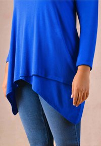 Blå långärmad topp med asymmetrisk fåll, tillverkad av mjukt tyg. Bärs över åtsittande blå jeans med en slät yta och utan synliga mönster.