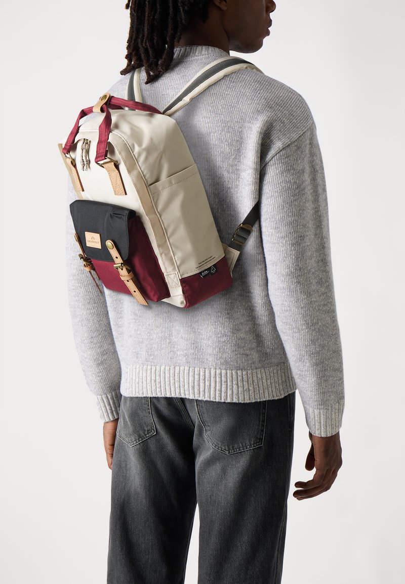 Sac à dos avec un corps beige, un fond bordeaux et un rabat noir, agrémenté de quincaillerie dorée et d'accents en cuir. Porté par-dessus un pull gris.
