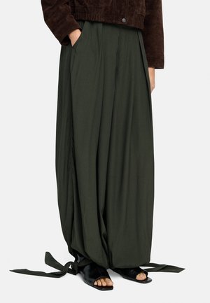 Luźne, ciemnozielone szerokie spodnie z związanymi nogawkami, w połączeniu z czarnymi sandałami z odkrytymi palcami i ciemnobrązową kurtką, ręce w kieszeniach.