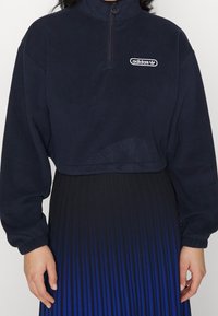 Person som bär en marinblå Adidas fleece med kvartslång dragkedja och logotyp på bröstet samt en högmidjad svart till blå gradient veckad kjol.