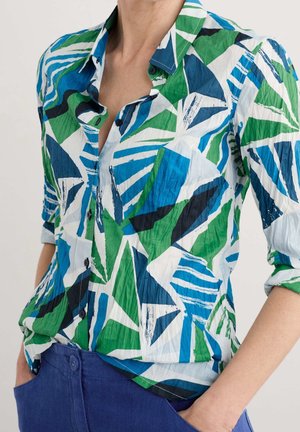 Kobieta ubrana w pomiętą koszulę z geometrycznymi wzorami w odcieniach niebieskiego, zieleni, bieli i czerni oraz niebieskie spodnie, trzymająca ręce w kieszeniach.