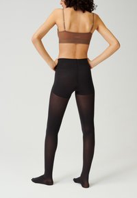 Collants opaques noirs avec une texture lisse, taille haute et finition mate, portés avec un bralette marron, mettant en avant un design épuré.