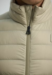 Giacca beige trapuntata con un tirante a forma di triangolo nero, indossata da una persona visibile dalla mascella alle spalle.