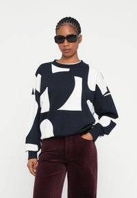 Sudadera de rayas en azul marino y blanco con un diseño abstracto, cuello redondo y puños de canalé, combinada con pantalones de pana burdeos. Gafas de sol negras.