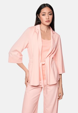 Woman with long dark hair wearing a light pink blazer with tied waist, matching pants, and a pink top against a plain light background.