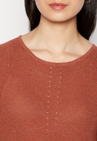 Rustfarbener Strickpullover mit rundem Halsausschnitt und vertikalen Reihen kleiner Löcher in der Mitte, mit einer weichen, strukturierten Oberfläche.
