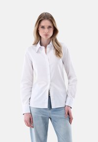 Camicia bianca con colletto classico, maniche lunghe e sottili dettagli di cucitura. Abbinata a jeans azzurri.