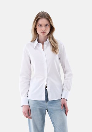 Camicia bianca con colletto classico, maniche lunghe e sottili dettagli di cucitura. Abbinata a jeans azzurri.