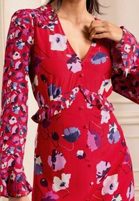 Vestido rojo con un patrón floral en púrpura y blanco, con un escote en V profundo, cintura con volantes y cierre de botones en la parte delantera. Textura de tela suave.