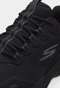 Μαύρο αθλητικό παπούτσι με πλεκτό επάνω μέρος, λογότυπο στην πλευρά και ανακλαστική ριγωτή θηλιά που διατρέχει τα μαύρα κορδόνια.