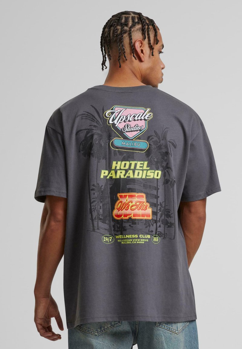 Grå oversized t-shirt med farverige grafik på bagsiden, herunder palmer, "Upscale Studios Malibu" og "Hotel Paradiso."
