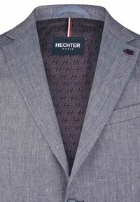 Grau strukturierter Blazer mit feinem Fischgrätenmuster, ausgestattet mit einem dunklen gemusterten Futter und einem Logotag. Knopf- und Taschen details sind sichtbar.