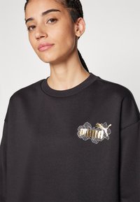 Felpa nera con un logo floreale ricamato in oro e bianco sul petto. Il tessuto sembra morbido, con una vestibilità comoda e un collo rotondo.