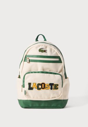 Sac à dos Lacoste en toile beige avec des accents verts. Équipé d'un logo brodé et du texte "LACOSTE" en multicolore, ainsi que de deux poches zippées.