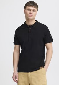 Černé polo tričko z bavlněné směsi s klasickým límečkem, třemi hnědými knoflíky a detailem božího štítku. Nosené s khaki šortkami.