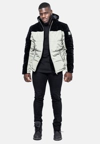 Vadderad jacka med ljusgrå kropp och svarta sammetsärmar, med dragkedjestängning, huva och framfickor, bärs över en svart topp.