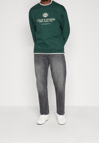 Person som bär en mörkgrön sweatshirt med texten "Legacy Manor", urblekta svarta jeans och vita sneakers, stående mot en enkel vit bakgrund.