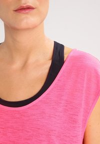 Svítivě růžové sportovní tričko s texturovaným povrchem a širokým výstřihem, vrstvené přes černý sportovní top. Krátké rukávy a ležérní design.