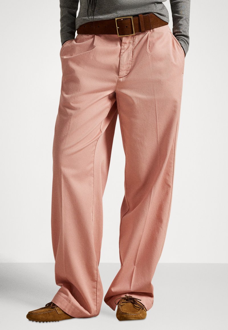 Jasnoróżowe spodnie o szerokich nogawkach wykonane z gładkiej tkaniny, z bocznymi kieszeniami i brązowym skórzanym paskiem, połączone z brązowymi zamszowymi butami.