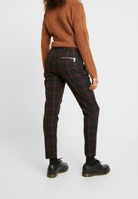 Svarta byxor med ett rött rutmönster, slim-fit design, dragkedjeficka baktill, kombinerat med en texturerad brun tröja och svarta skor.