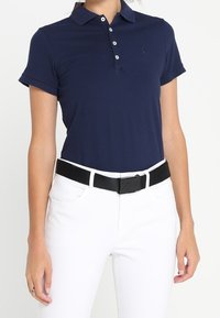 Mörkblå pikétröja med korta ärmar, ribbad krage, placket med tre knappar och en broderad logotyp, kombinerad med vita byxor och ett svart bälte.