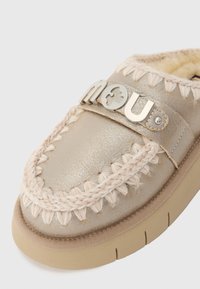Metallisk gullmoccasin med teksturert finish, flettet tauakent og en sølv logo stropp. Lys brun såle med ribbet design for grep.