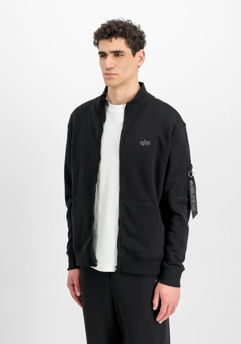 Jeune homme aux cheveux bouclés portant une veste noire zippée avec un petit logo et une chemise blanche en dessous, debout devant un fond blanc uni.
