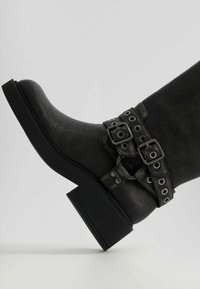 Botín de cuero negro con punta redondeada, tacón grueso de bloque y doble correa con hebillas y ojales de metal. Acabado texturizado.