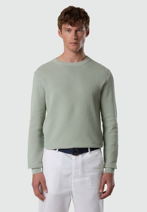 Junger Mann trägt einen hellgrünen, strukturierten Pullover, weiße Hose und einen marineblauen Gürtel, steht vor einem schlichten grauen Hintergrund.