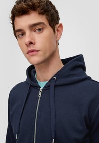 Sudadera con cremallera en color azul marino y capucha con cordón. Textura suave, cremallera plateada y camiseta interior azul clara visible debajo.