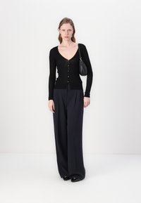 Schwarze gerippte Strickjacke mit V-Ausschnitt und Knöpfen, kombiniert mit navyfarbener weitem Hose. Das Model hält eine kleine schwarze Handtasche. Insgesamt glatte Textur.