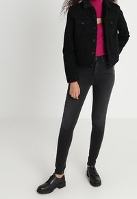 Persona vestida con chaqueta de mezclilla negra, suéter de cuello alto magenta, jeans skinny negros y zapatos negros con cordones, de pie contra un fondo liso.