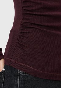 Burgunderfarbener Ripp-Top mit gerafftem Seitendetail, das ein tailliertes Design und kontrastierende Nähte aufweist. Kombiniert mit schwarzen, strukturierten Jeans.