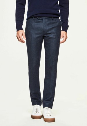 Mand iført marineblå slim-fit bukser, mørkeblå sweater og hvide sneakers med brune såler, stående mod en ensartet lys baggrund.