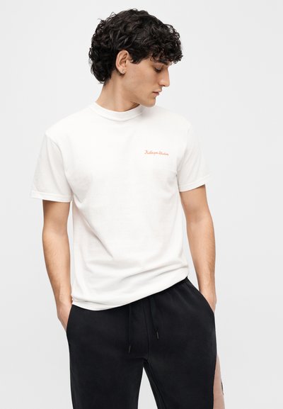 Λευκό βαμβακερό T-shirt με στρογγυλή λαιμόκοψη, διακοσμημένο με κεντημένο κόκκινο κείμενο στο στήθος. Σε συνδυασμό με μαύρα παντελόνια φόρμας, εμφανίζεται σε πλάγια όψη.