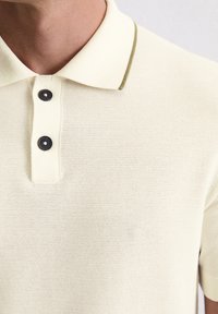 Kremowa koszulka polo z teksturowanej tkaniny, czarnymi guzikami i oliwkowym akcentem na kołnierzyku. Zbliżenie na cechy wzornictwa.