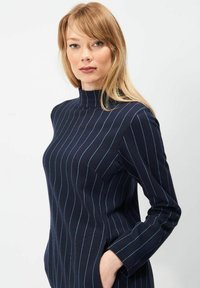 Marinekjole med lodrette lyseblå striber, høj hals, lange ærmer og sidelommer. Blødt stof med en tætsiddende silhuet.