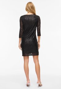 Woman wearing a black, sequined, knee-length dress with three-quarter sleeves, standing facing away, paired with white high-heeled sandals.