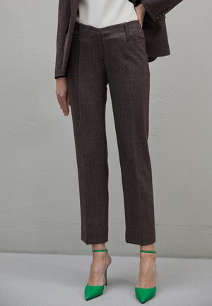 Une personne portant un pantalon marron sur mesure, un blazer assorti, un haut blanc et des chaussures à talons hauts à bride de cheville vert fluo se tenant contre un mur gris.