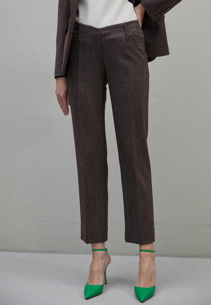 Άτομο που φοράει καφέ κομμένα παντελόνια, ταιριαστό σακάκι, λευκό τοπ και φωτεινά πράσινα ψηλοτάκουνα παπούτσια με ανακρουστικό λουράκι, στέκεται μπροστά σε γκρίζο τοίχο.