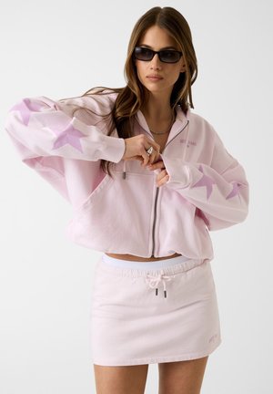 Donna che indossa occhiali da sole neri, felpa rosa chiaro con cerniera e stelle sulle maniche, e gonna rosa chiaro abbinata con coulisse in vita.