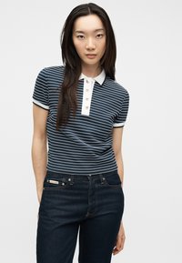 Νεαρή γυναίκα με μακριά σκούρα μαλλιά, που φοράει μπλε ναυτικό και λευκό ριγέ κοντομάνικο πόλο μπλουζάκι και σκούρα μπλε τζιν, μπροστά σε απλό φόντο.