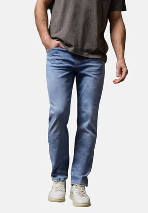 Uomo che indossa jeans azzurri, sneakers bianche e una t-shirt grigio scuro, camminando con una mano in tasca su sfondo bianco.