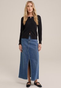 Zwarte gebreide cardigan met knopen aan de voorkant, gedragen met een lange spijkerrok met een split aan de voorkant. Model draagt zwarte platte schoenen.