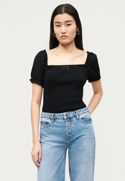 Kvinne med langt mørkt hår som har på seg gull hoops-øreringer, en svart topp med firkantet hals og pufferm, og lyseblå høytlivsjeans mot en ensfarget bakgrunn.