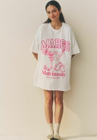 Vit oversized t-shirt klänning med rosa grafisk text "MARGS" tillsammans med en cocktail och ringar, tillverkad av mjukt bomullstyg.