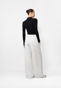 Žena s účesom v drdole, oblečená v čiernom top s dlhými rukávmi a voľných svetlošedých teplákoch, stojí na bielom pozadí.