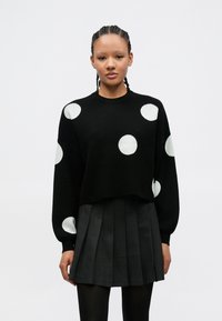 Suéter preto de malha com grandes bolinhas brancas, com decote redondo e ombros caídos, combinado com uma saia preta plissada.