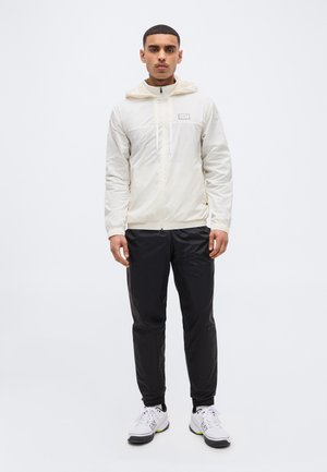 Νεαρός άνδρας όρθιος, φορώντας ανοιχτό μπεζ φούτερ με κουκούλα, μαύρο παντελόνι και λευκά αθλητικά παπούτσια, μπροστά σε απλό λευκό φόντο.